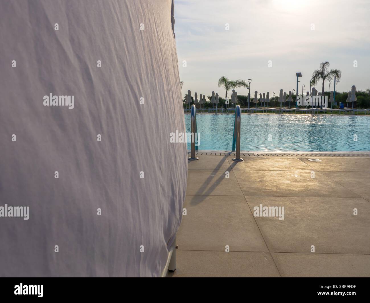 Blick von hinter dem Sonnenschirm mit weißen Netzvorhängen über einen leeren Pool mit Liegestühlen, geschlossenen Sonnenschirmen und Palmen im tropischen Resort Stockfoto