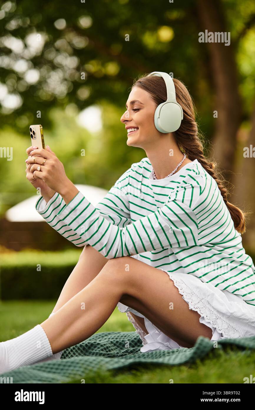Eine lächelnde Frau sitzt auf einer Picknickdecke, genießt Musik und macht Fotos im Park. Stockfoto