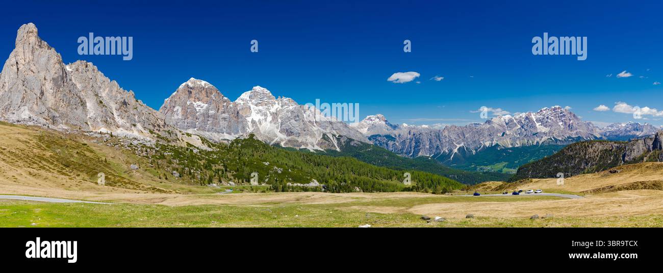 A sweeping panoramic view from Passo di Giau, showcasing rugged Dolomite peaks, verdant valleys, and a clear blue sky, with a winding road below. Stockfoto