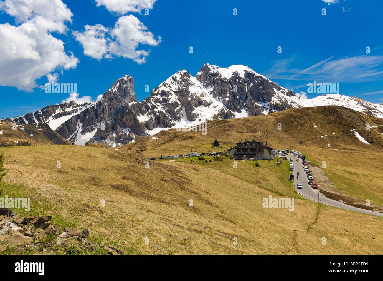 The winding road of Passo di Giau leads to a mountain lodge, with the snow-capped peak of Antelao dominating the background under a vibrant blue sky. Stockfoto