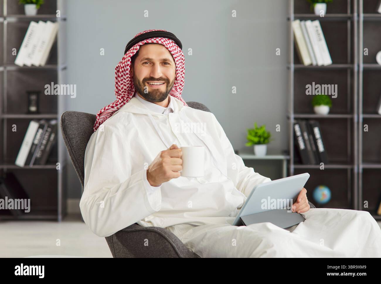 Arabischer Mann, der während der Fernarbeit eine Kaffeepause macht, posiert mit einer Tasse Kaffee, einem digitalen Tablet Stockfoto