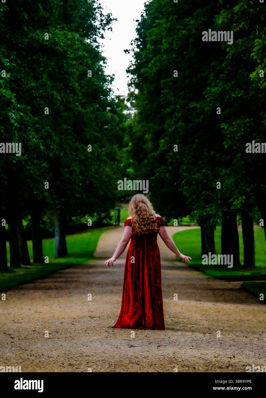 Eine Frau in rotem Kleid steht auf einem Weg in einem Park, mit gestreckten Armen. Stockfoto