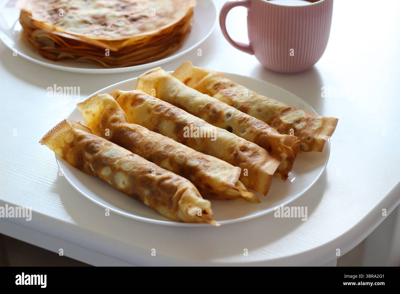 Gerollte Crepes mit Hüttenkäsefüllung auf weißem Teller. Rosafarbene Tasse im Hintergrund mit weichem Fokus. Gemütliches Frühstück oder leichte Dessertidee Stockfoto