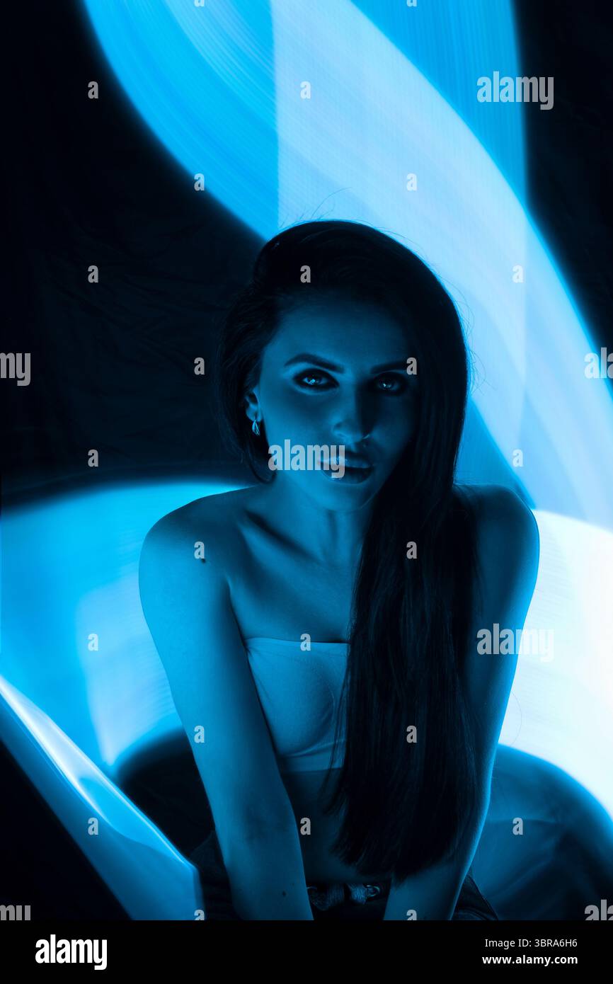 Frau posiert vor einem Hintergrund von wirbelndem blauem Licht, hebt ihre Eigenschaften hervor und erzeugt einen ätherischen Effekt. Die künstlerische Verwendung von Licht verleiht der Komposition Tiefe und Intrige Stockfoto