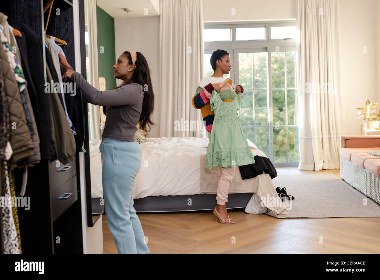 Verschiedene Freundinnen wählen Kleidung aus einem offenen Kleiderschrank in einem hellen Schlafzimmer mit grünem Kleid aus Stockfoto