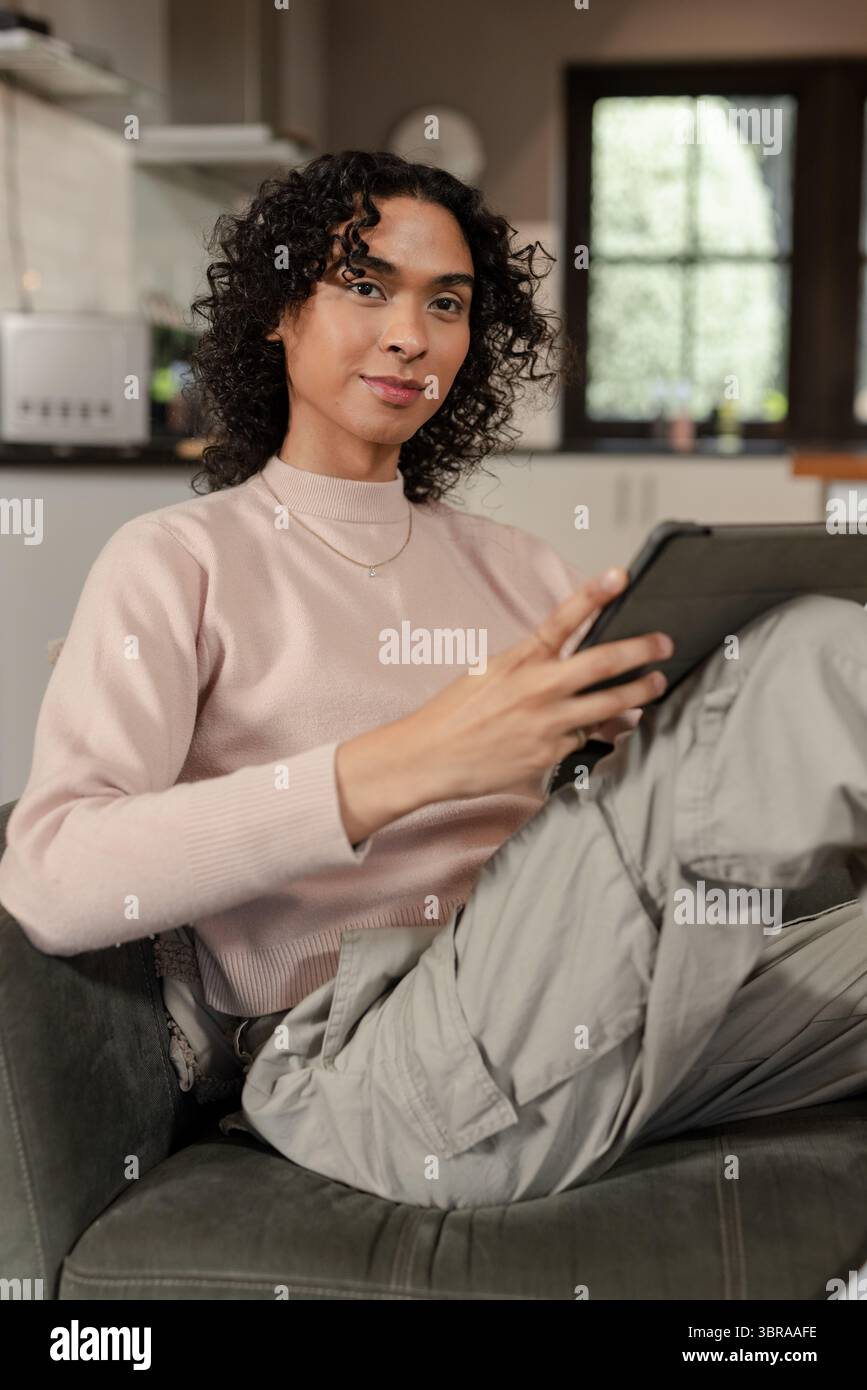 Nicht binäre Person, die auf dem Sofa sitzt, einen Pullover trägt und in der Küche ein Tablet in der Nähe einer Kaffeemaschine hält Stockfoto