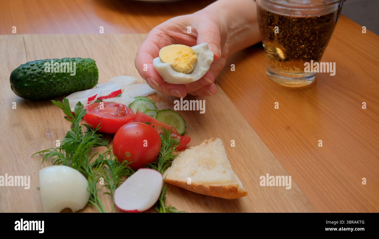 Eine Hand hält ein gekochtes Ei, während frisches Gemüse es auf einem Holzbrett umgibt. Stockfoto