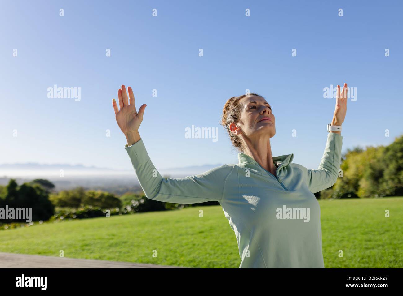 Frau, die sich im Park streckt, hebt die Arme nach oben und trägt einen grünen, weißen Fitness-Tracker mit Viertelreißverschluss Stockfoto