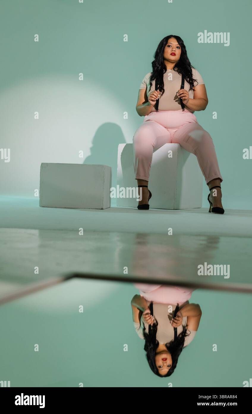 MittelErwachsene Frau mit Hosenträgern, die auf weißem Würfel auf dem Boden vor pastellgrünem Hintergrund sitzt Stockfoto