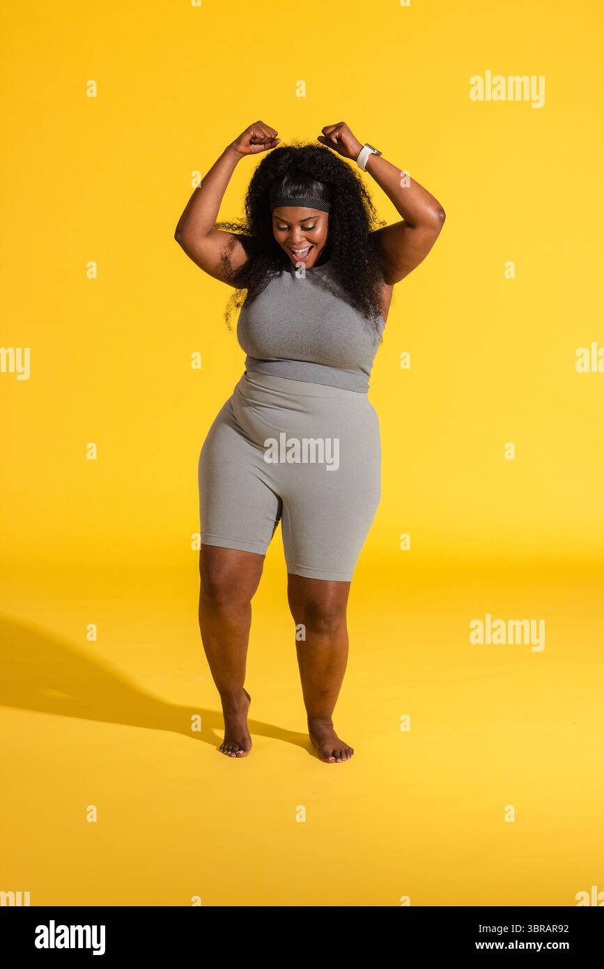 Eine Frau in Übergröße, die auf gelbem Boden lächelnd Fäuste in Sportbekleidung und Smartwatch hebt, die Schatten wirft Stockfoto