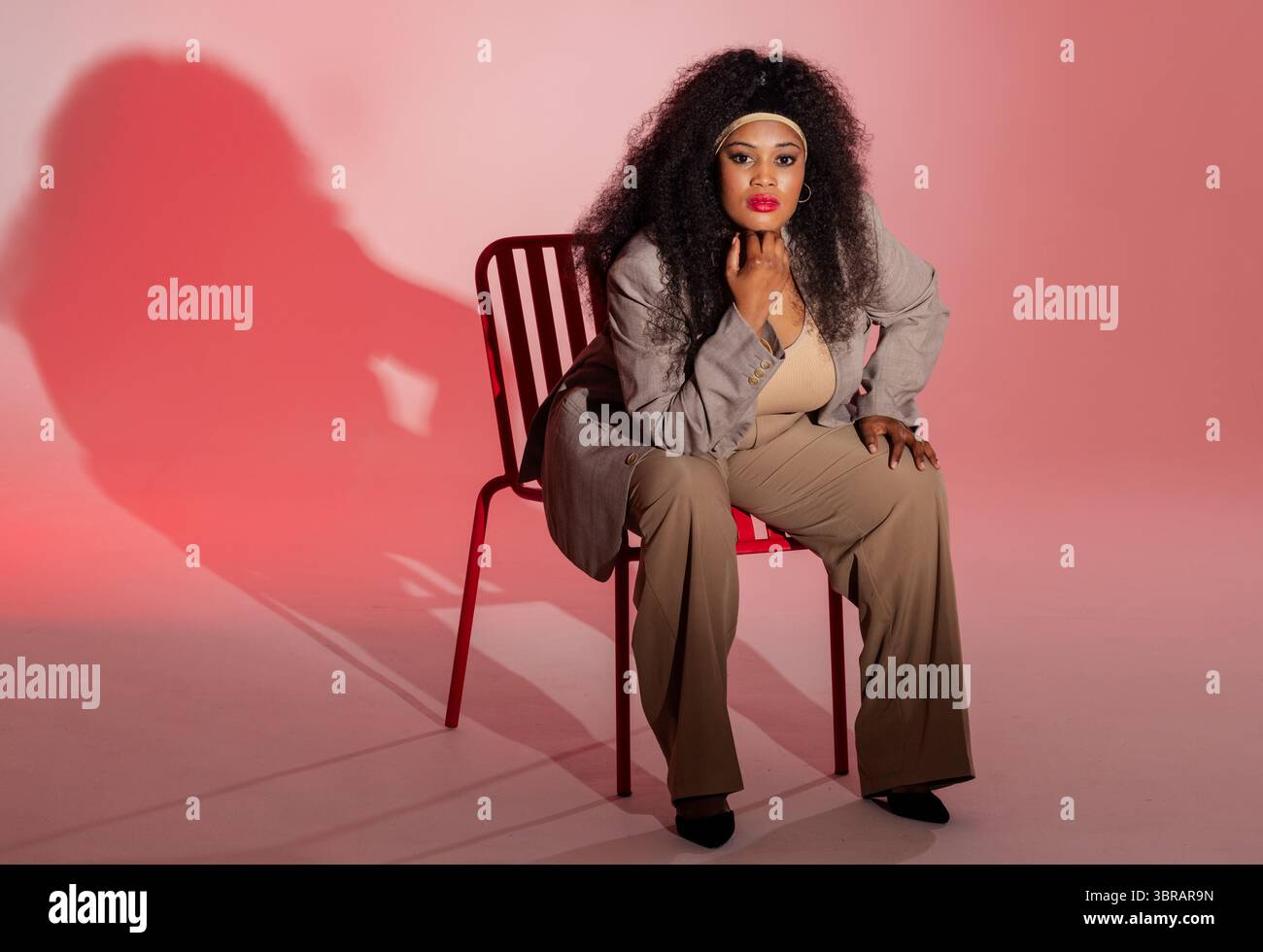 Afroamerikanerin im beigefarbenen Anzug, die sich in rosa-rotem Studio auf rotem Stuhl nach vorne lehnt Stockfoto