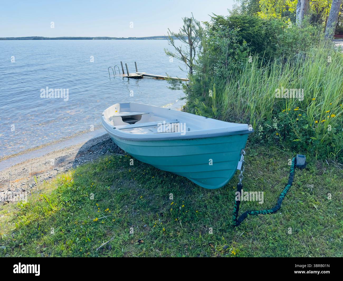 Ein kleines blaublaues Boot ruht an einem grasbewachsenen Ufer mit Blick auf einen ruhigen See an einem sonnigen Tag. - Smartphone-aufgenommenes Stockfoto