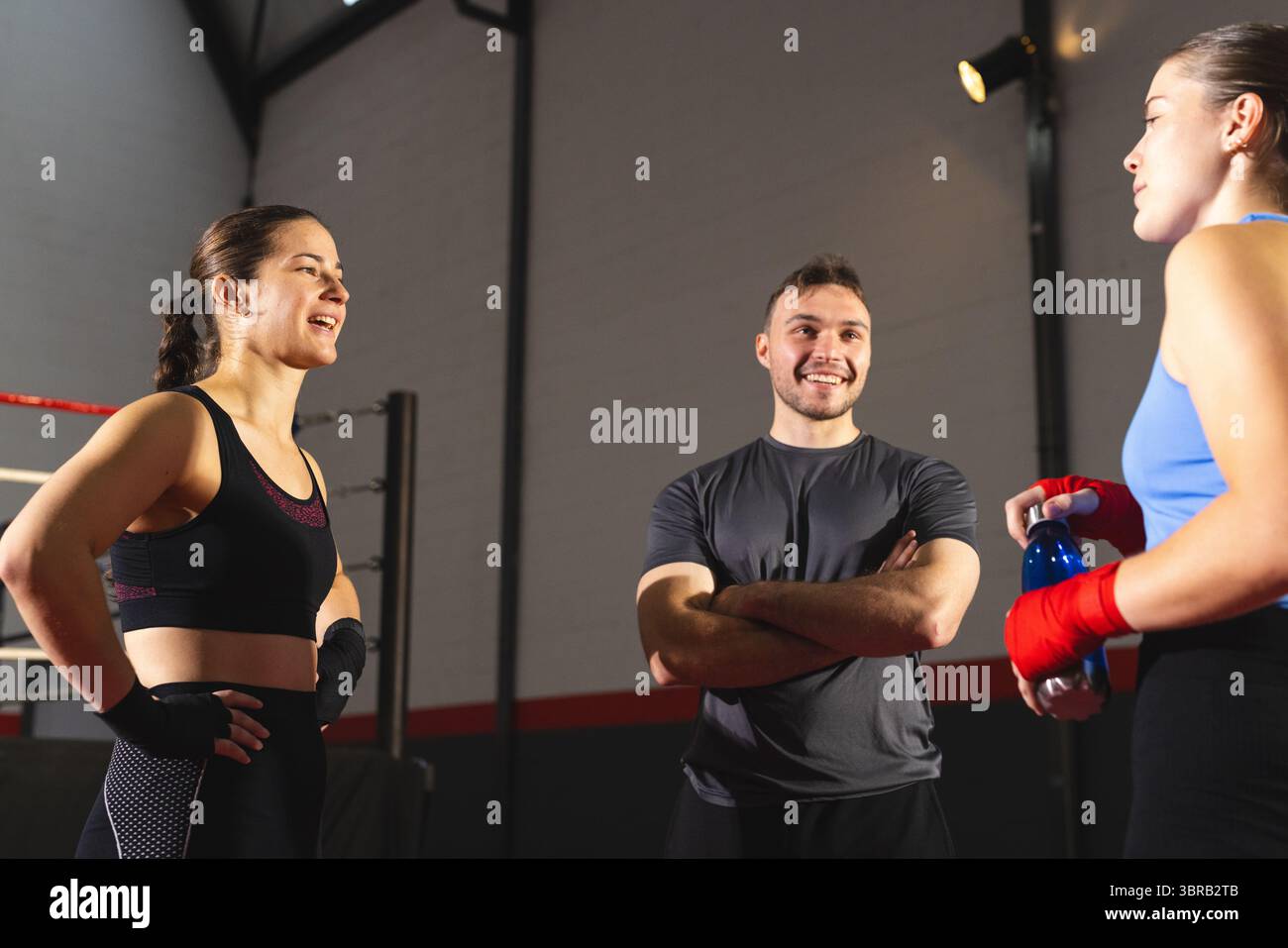 Männlicher Trainer, der zwei weibliche Auszubildende trainiert, die am Boxring eine Wasserflasche unter den Deckenleuchten halten Stockfoto