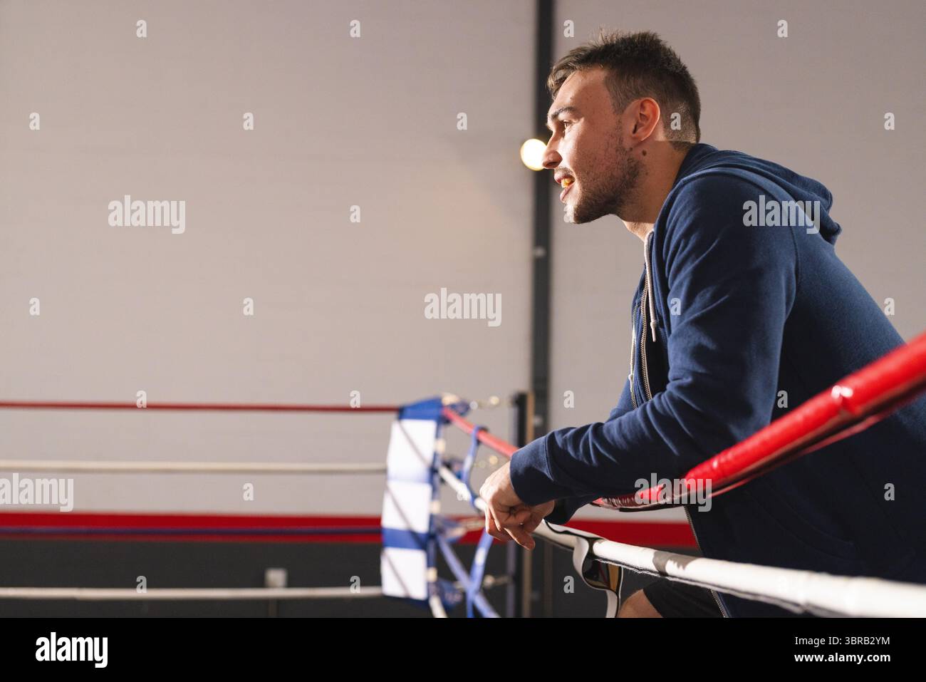 Mann lehnt sich an Boxringseile, trägt blauen Hoodie und schwarze Shorts im Fitnessstudio, Kopierraum Stockfoto