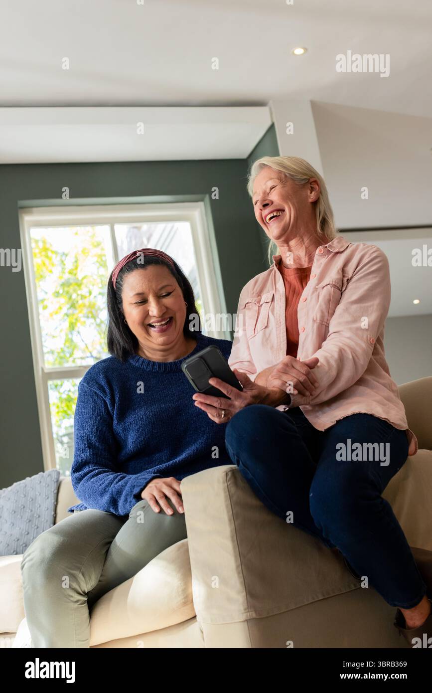 Verschiedene Freundinnen lachen und teilen ihr Smartphone, während sie im Wohnzimmer auf dem beigefarbenen Sofa sitzen Stockfoto