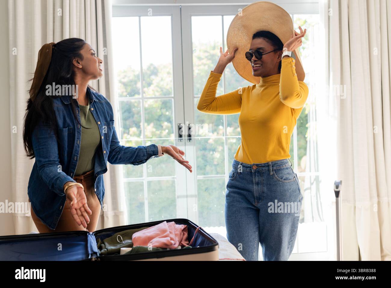 Verschiedene Freundinnen probierten Strohhut und Sonnenbrille neben dem offenen Koffer im hellen Schlafzimmer an Stockfoto