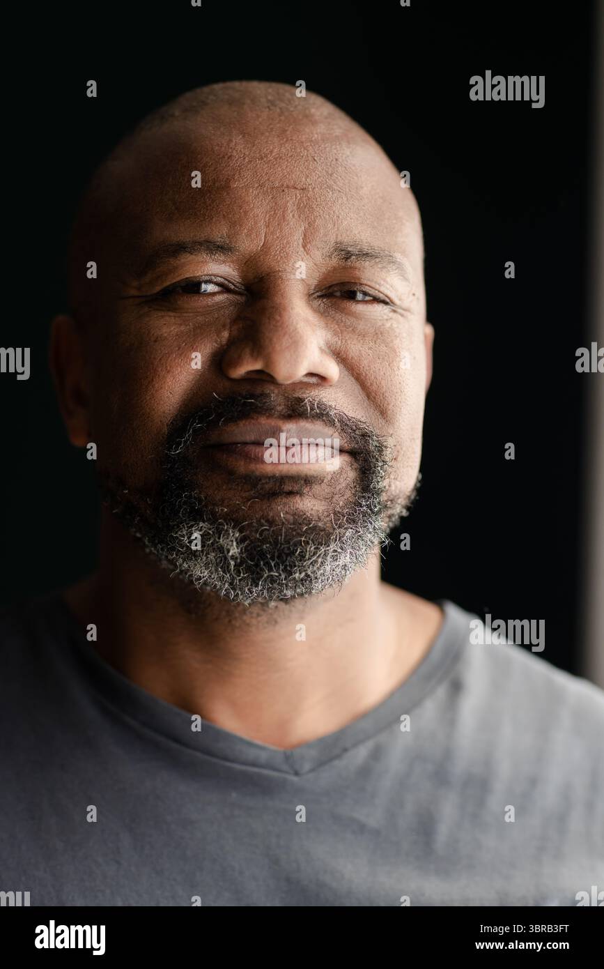 Ranghoher Afroamerikaner lächelnd und posierend im Zimmer mit dunkler Kulisse und Fensterlicht Stockfoto
