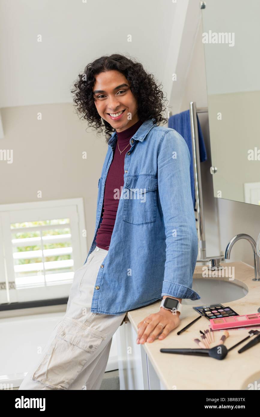 Nicht-binärer Erwachsener, der sich auf die Ablage im Badezimmer neben einem modernen Waschbecken lehnt und lächelt, während er Make-up-Pinsel arrangiert Stockfoto