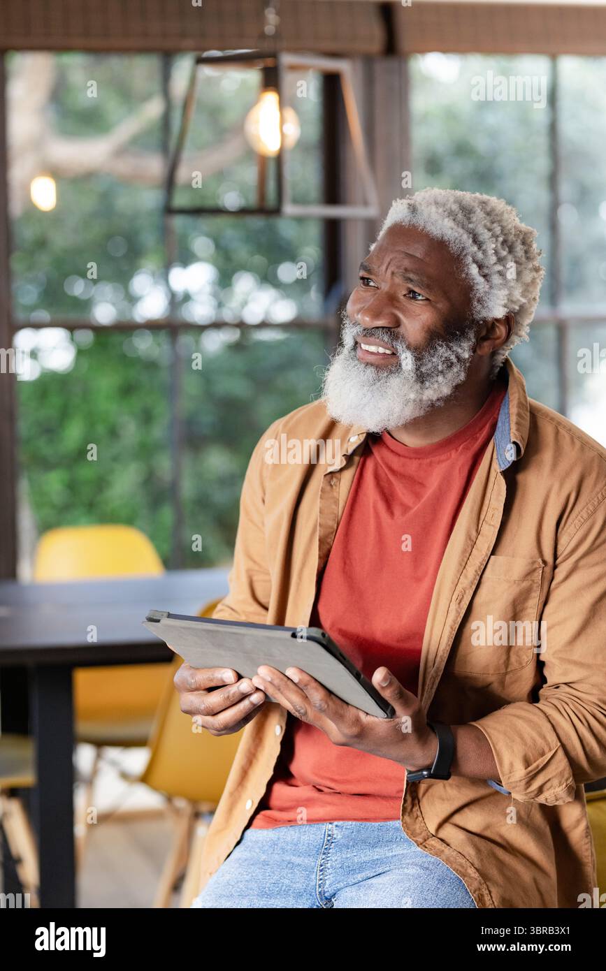 Ranghoher afroamerikanischer Mann, der am Tisch sitzt und eine Tablette unter einer Metallleuchte in der Nähe des Fensters hält Stockfoto