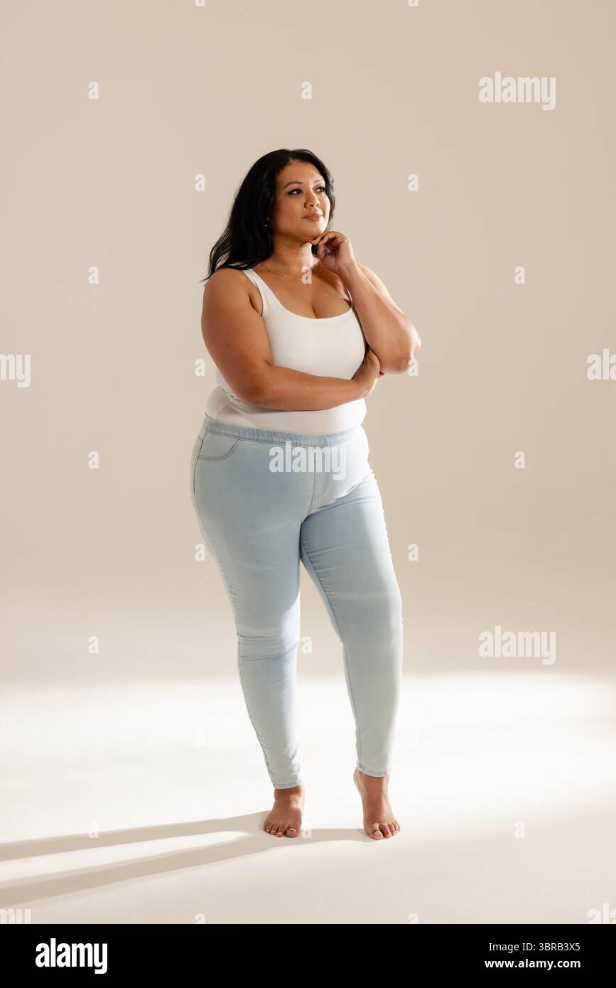 Frau, die Arme überkreuzte Hand auf dem Kinn in einem Studio in neutralem Ton trägt, in weißem Oberteil und Jeans Stockfoto