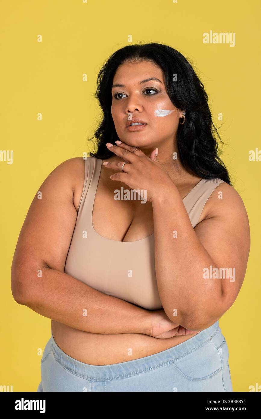 Frau, die mit der Hand auf dem Kinn im gelben Atelier steht, zeigt Creme-Streifen auf Wange und Ring Stockfoto