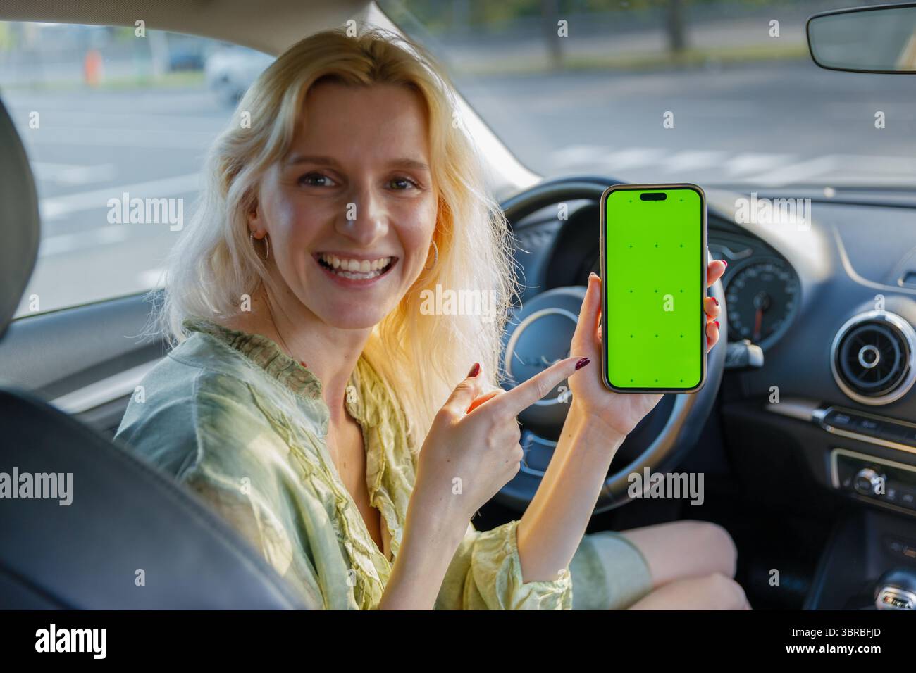 Eine lächelnde Frau im Auto hält ein Telefon mit einem hellen grünen Bildschirm hoch, zeigt fröhlich darauf und zeigt moderne Technologie und Konnektivität Stockfoto