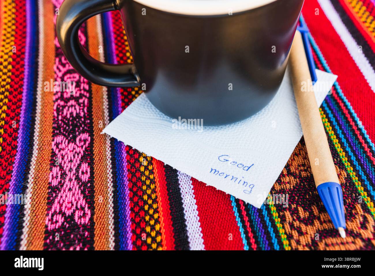Kaffeetasse mit einer Nachricht, die einen guten Morgen auf einem farbenfrohen Tisch mit Inka-Gewebe sagt. Stockfoto