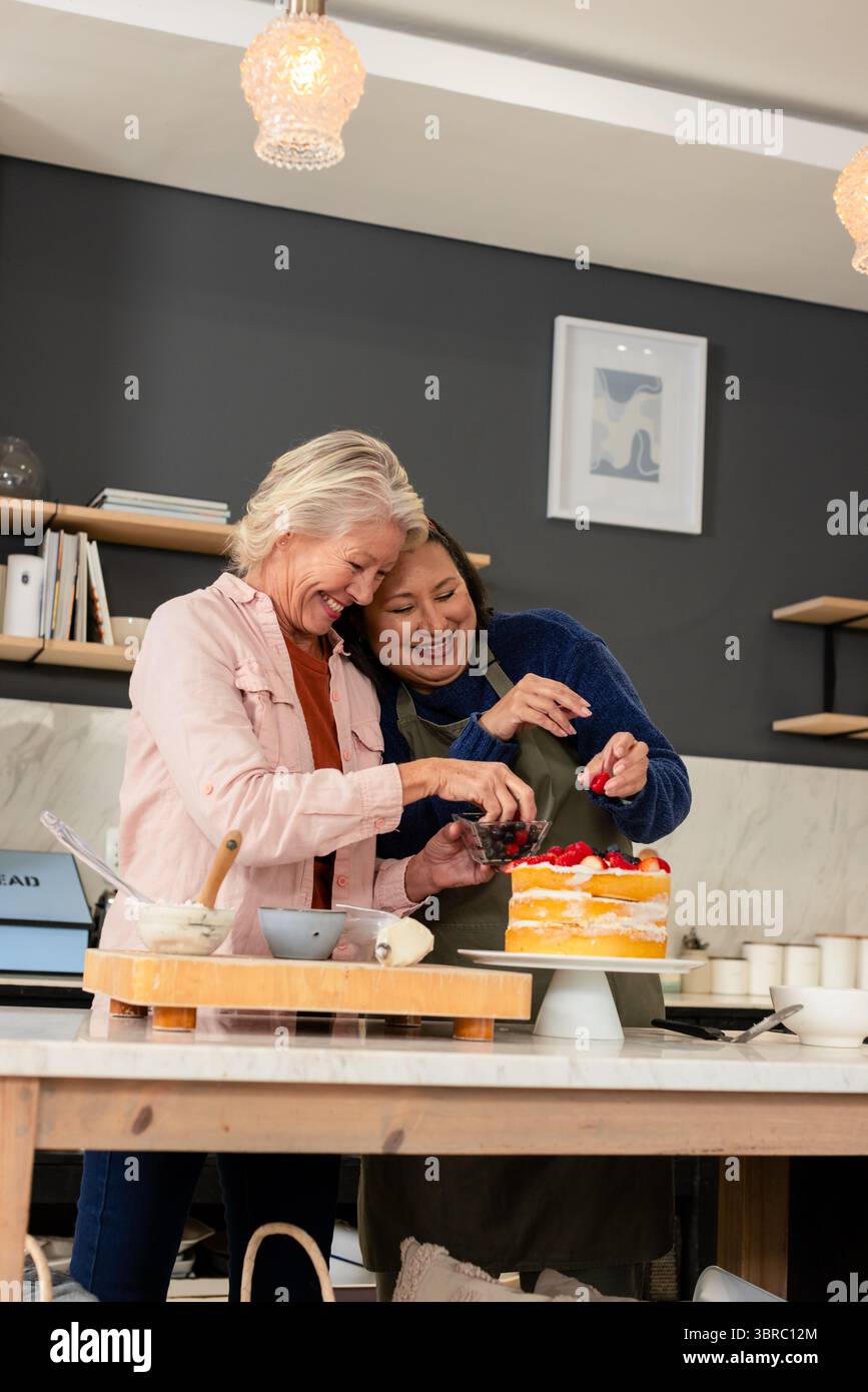 Ältere Freundinnen dekorieren mehrschichtigen Biskuitkuchen auf der Kücheninsel zu Hause mit Beeren Stockfoto