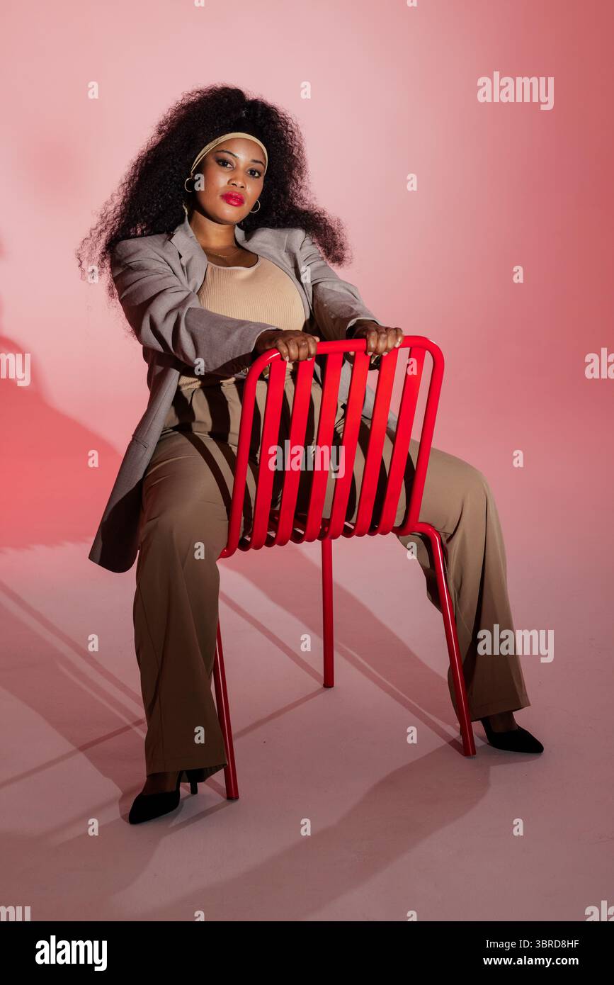 Afroamerikanerin, die einen Blazer trägt, sitzt rückwärts auf rotem Stuhl im rosa Studio Stockfoto