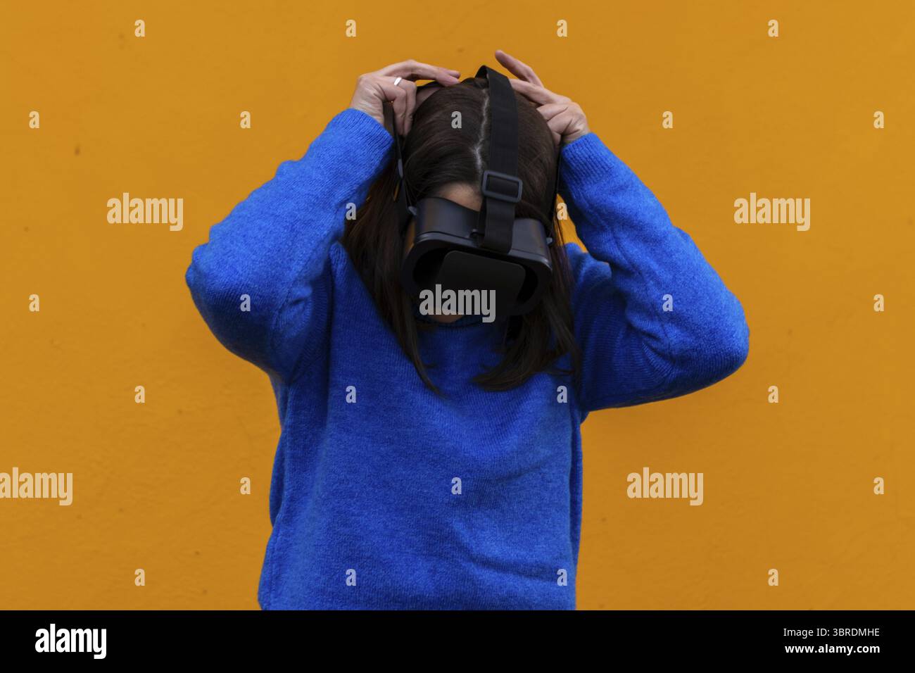 Eine junge Frau in einem Jugendoutfit benutzt eine Virtual-Reality-Brille. Eine Brünette in Blau auf gelbem Hintergrund bringt ihr ein vr-Gadget auf den Kopf. Eintauchen in ein VI Stockfoto