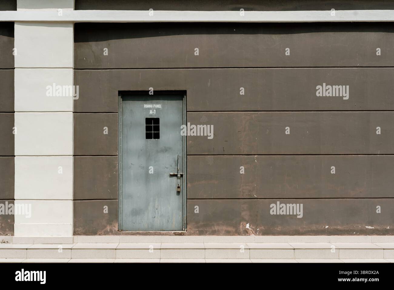 Minimalistisches architektonisches Detail einer Industrietür für einen Panelraum („RUANG PANEL A-1“) an einer braun-weißen Paneelwand mit Stufen unten. Stockfoto