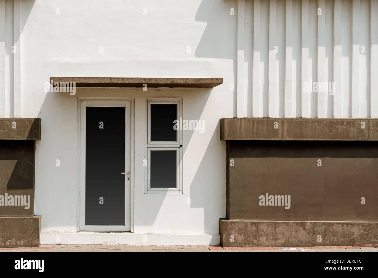 Minimalistische Fassade eines modernen Gebäudes mit einer weißen kannelierten Wand, braunen Akzenten, einer Tür und einem Fenster. Starke geometrische Linien und Schatten. Stockfoto
