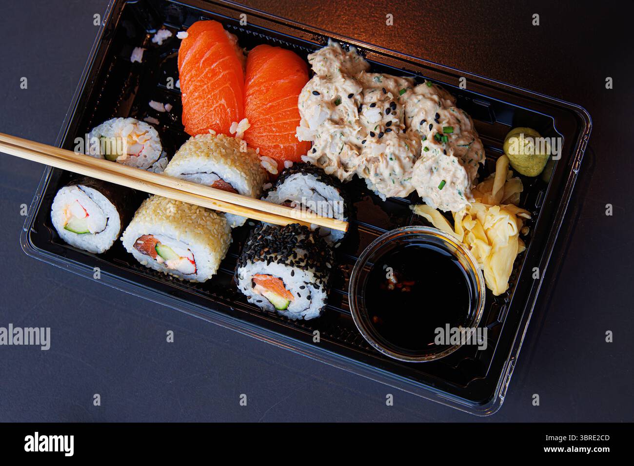 Ein Sushi-Essen zum Mitnehmen in einem schwarzen Plastiktablett mit Lachsnigiri, Maki-Brötchen, Ingwer, Wasabi und Sojasauce mit Essstäbchen über dem Tablett. Selektiv f Stockfoto