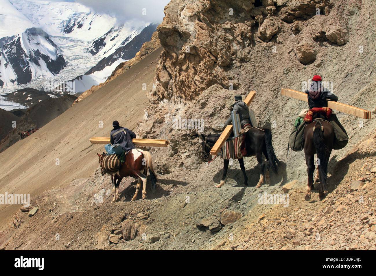Drei Träger zu Pferd tragen Lasten entlang des zerklüfteten Weges zu den Lenin Peak Camps. Raue Berglandschaft, Felsen und schneebedeckte Gipfel in der Ferne. Stockfoto