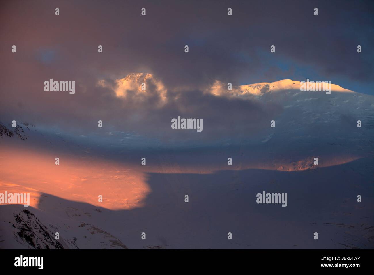 Der Lenin Peak leuchtet bei Sonnenuntergang durch einen Bruch der Gewitterwolken. Eisbedeckte Gipfel, die in Gold zwischen Schatten beleuchtet sind – eine dramatische, beeindruckende Bergszene. Stockfoto