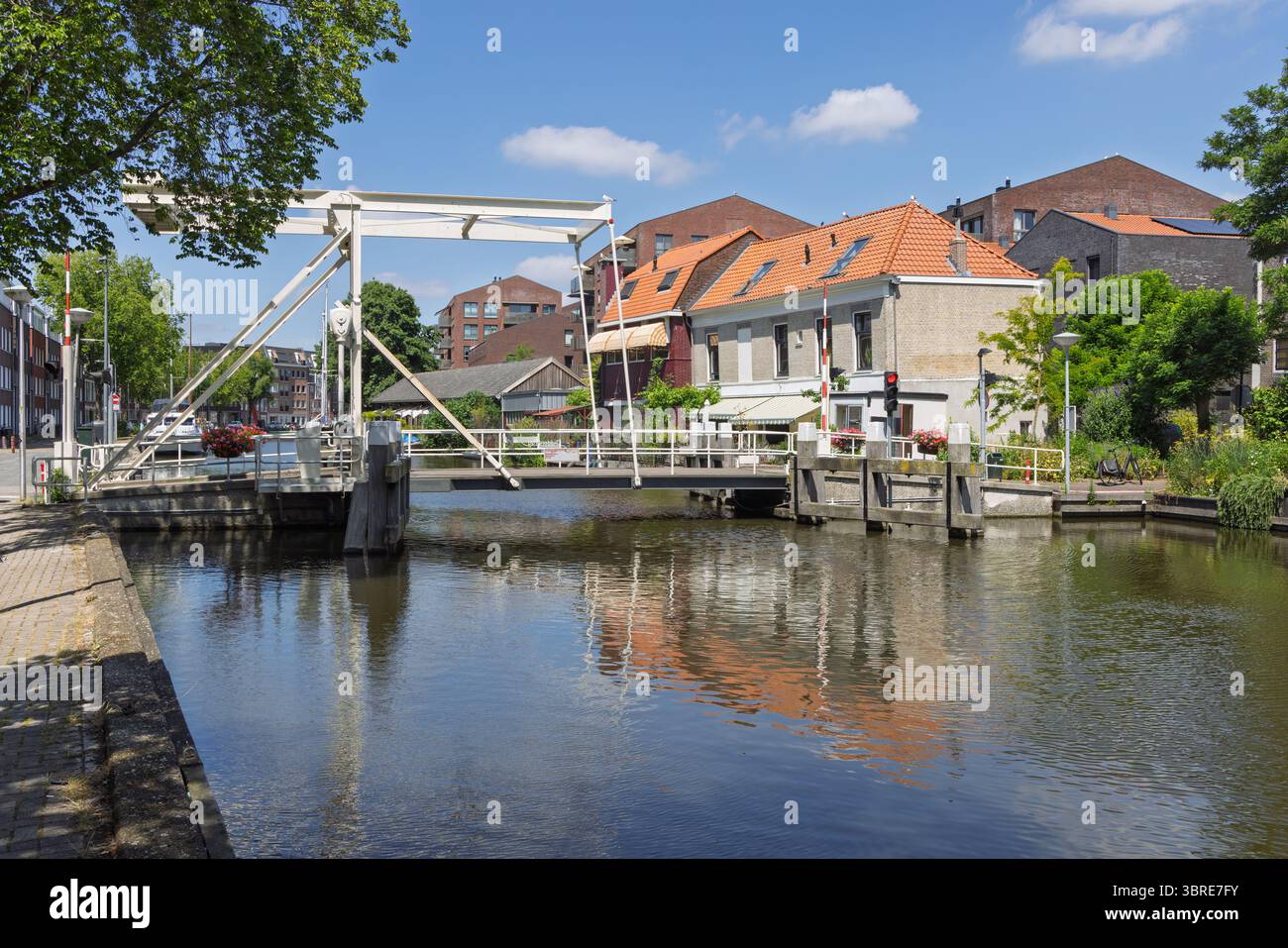 Eine bezaubernde Zugbrücke in Gouda überspannt einen ruhigen niederländischen Kanal, der an einem sonnigen Tag von malerischen historischen Gebäuden und üppigem Grün umgeben ist. Stockfoto