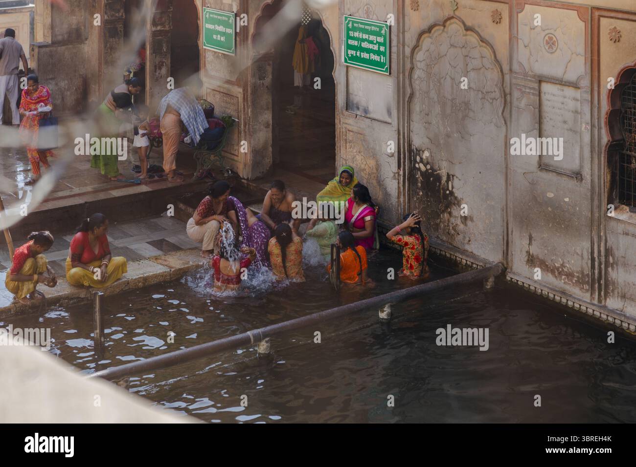 Jaipur, Indien - 24. Juni 2025: Blick auf Frauen im ruhigen Wasser des heiligen Kunds des Galtaji-Tempels, umgeben von verwitterten Steinmauern und lebhaften Saris. Stockfoto