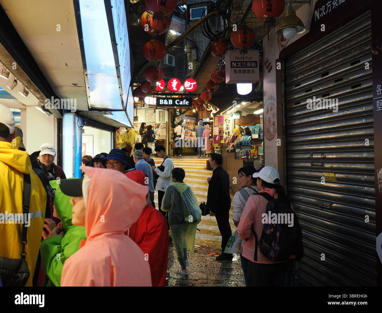 Jiufen, Taiwan - 8. November 2024 - Touristen in Regenkleidung und mit Sonnenschirmen steigen in eine geneigte, nasse Gasse der Jiufen Old Street, die von rotem Langhang beleuchtet wird Stockfoto