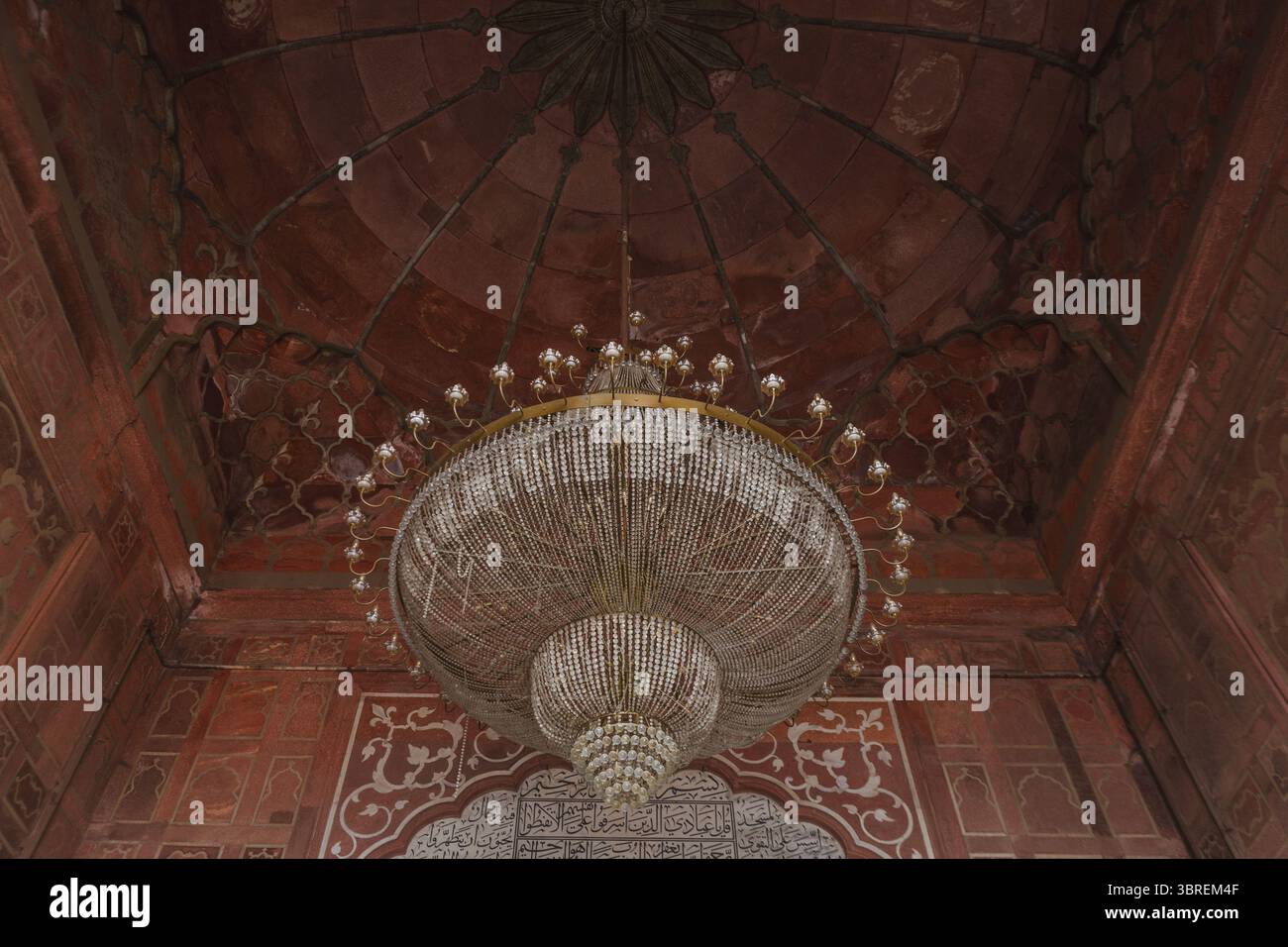 Blick auf einen großen Kronleuchter, der an einer rot verzierten Decke hängt und einen sanften Glanz auf die strukturierten Wände in Neu-Delhi, Delhi Division, Indien ausstrahlt. Stockfoto
