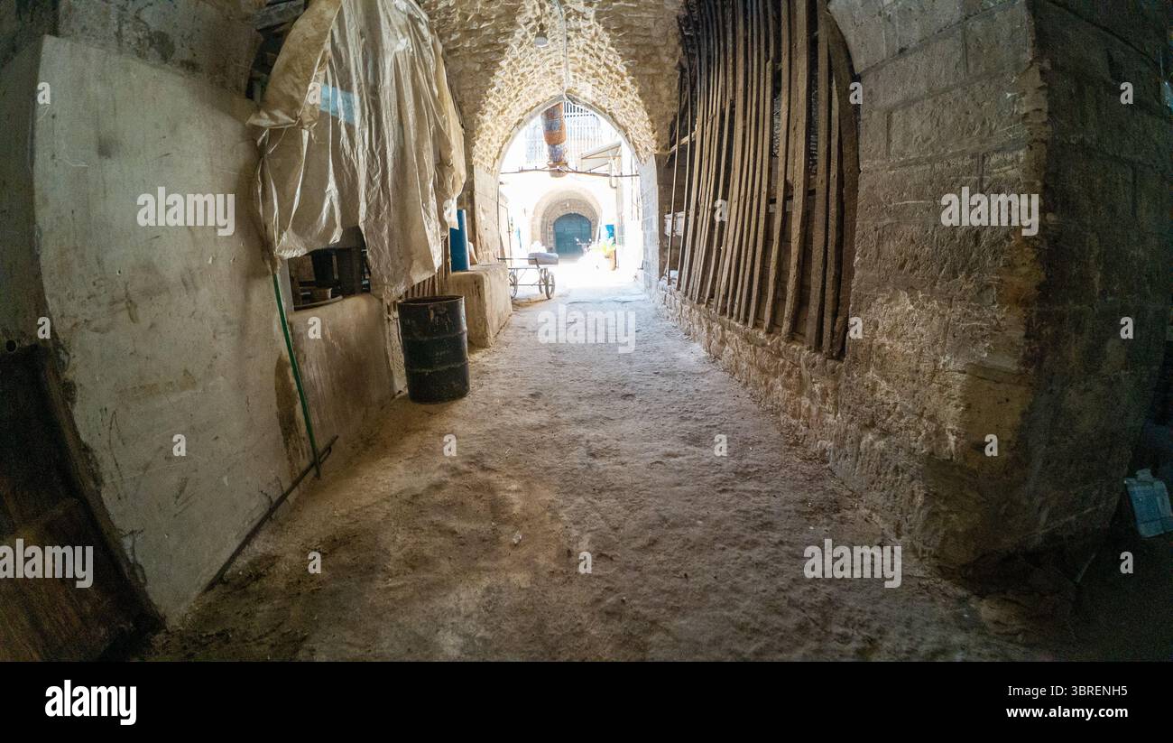 Ein schmaler, staubiger Korridor mit Steinmauern und einer Bogendecke, der zu einem hellen Eingang führt. Der Boden ist uneben und es gibt Baumaterialien Stockfoto