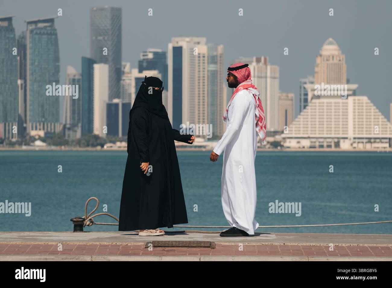 Ein Paar steht am Wasser und genießt das sonnige Wetter. Ein Mann und eine Frau in traditioneller arabischer Kleidung gehen am Damm entlang, gegen den Hintereingang Stockfoto