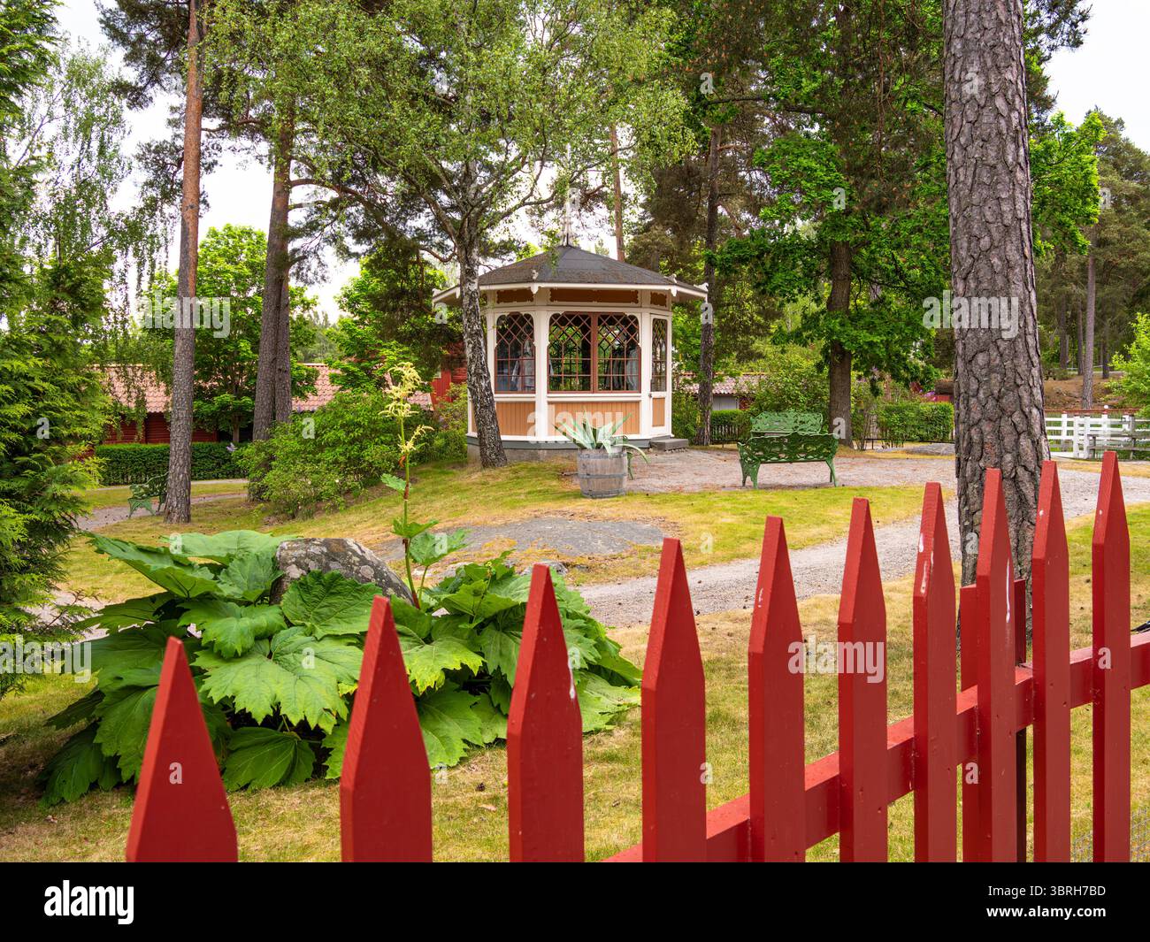 Sodertalje, Schweden - 6. Juni 2025: Eine bezaubernde Gartenszene mit einem Pavillon umgeben von üppigem Grün und einem roten Pfahlzaun. Der Pavillon ist lackiert Stockfoto
