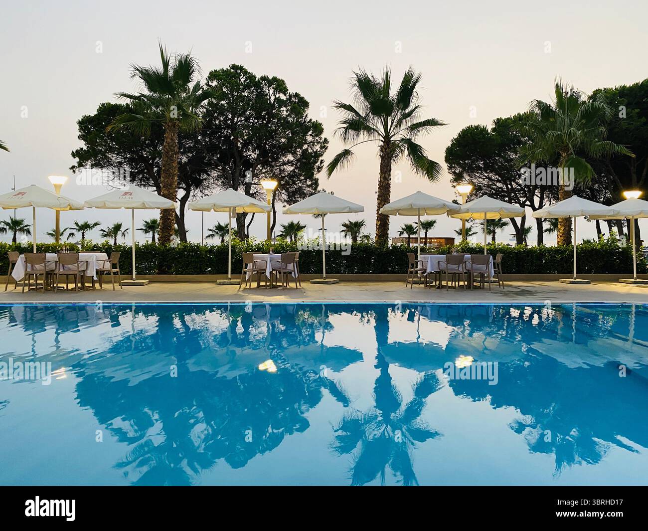 Der Poolbereich des Resorts oder des Hotels scheint in der Abenddämmerung oder am frühen Abend zu sein. Im Vordergrund spiegelt ein großer, klar blauer Swimmingpool die Atmosphäre wider Stockfoto