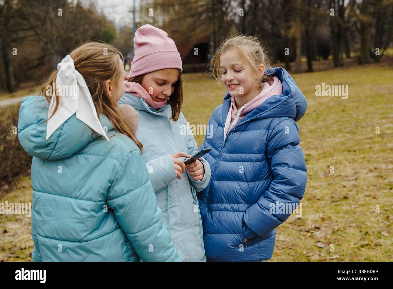 Teenagerinnen in Jacken und Jeans schauen Smartphone und hören Musik-Player im Winter-Stadtpark. Outdoor-Aktivitäten verbinden Kinder, bauen Freundschaft auf, s Stockfoto