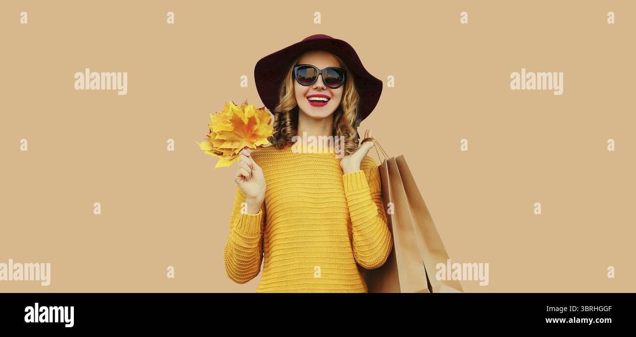 Herbstfarben-Outfit, Porträt einer schönen jungen Frau mit Einkaufstasche hält gelbe Ahornblätter tragen Hut, Strickpullover auf beigem Studio Stockfoto