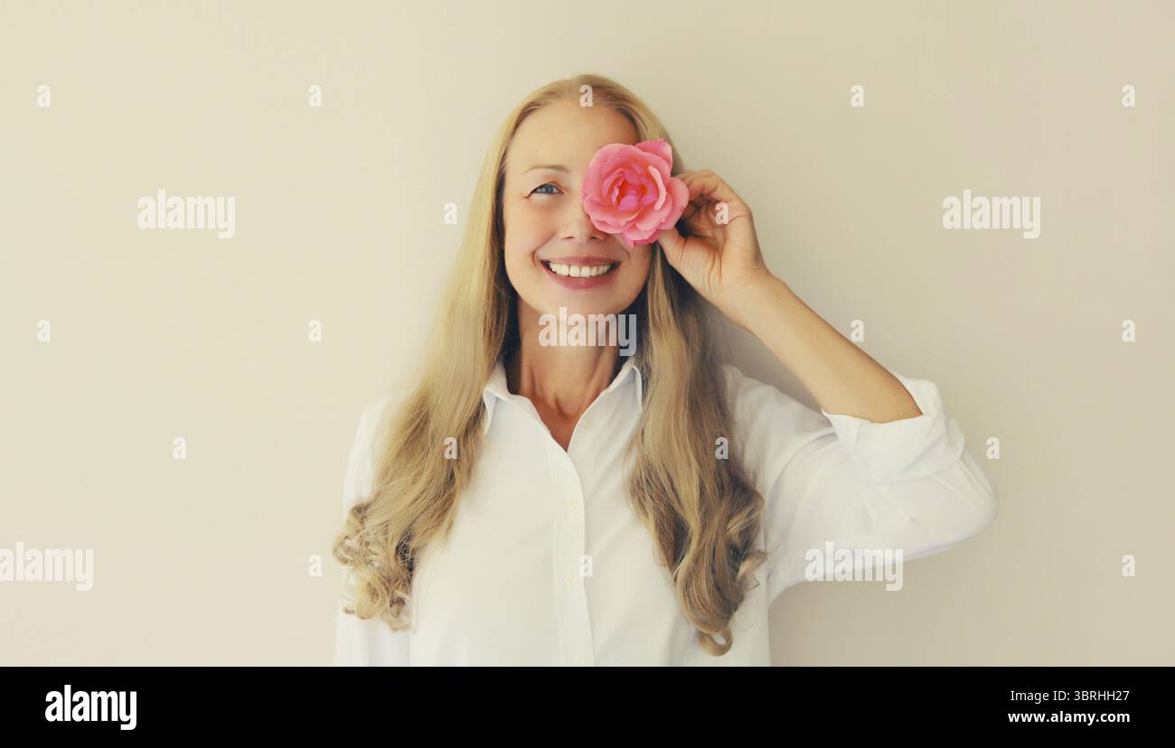 Sommerporträt einer glücklichen, lächelnden Frau mittleren Alters, die ihre Augen mit rosa Rosenblüten auf Studiohintergrund bedeckt Stockfoto