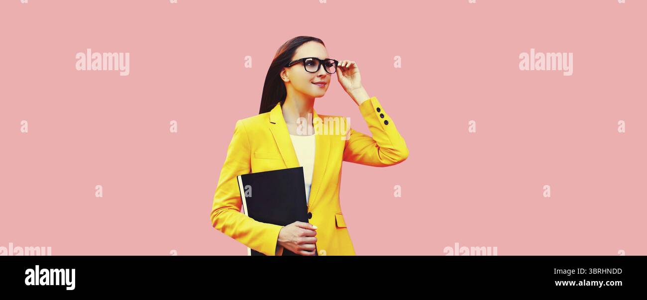 Porträt der eleganten jungen Frau mit Ordner, der wegschaut, Brille trägt, gelber Business-Anzug auf rosa Studiohintergrund Stockfoto