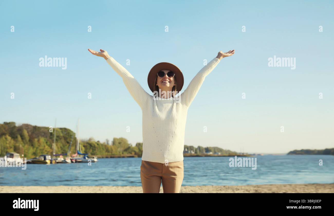Glückliche lächelnde Reiserin hebt ihre Hände am Strand, genießt warmes Wetter, trägt Hut, weißen Pullover, steht an der Küste, blauer Himmel, Naturbao Stockfoto