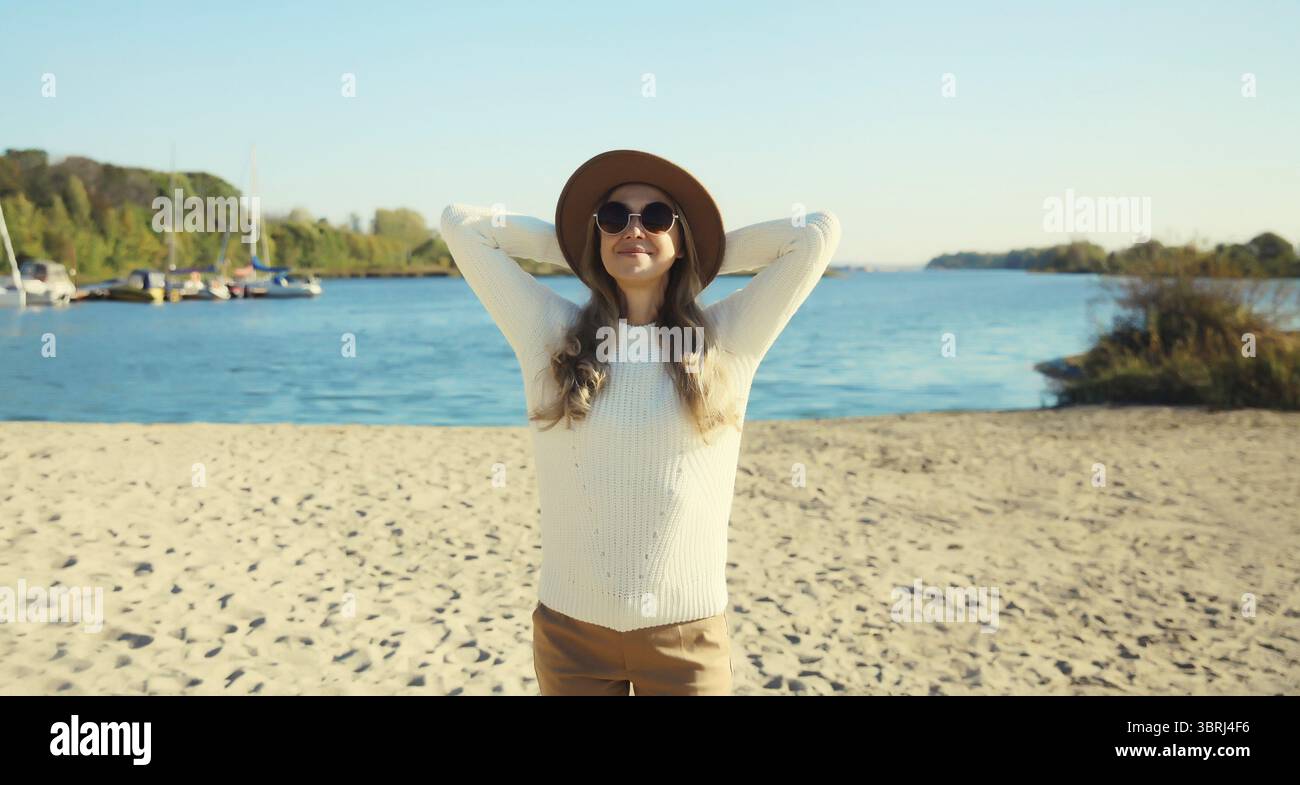 Glückliche lächelnde Reiserin hebt ihre Hände am Strand, genießt warmes Wetter, trägt Hut, weißen Pullover, steht an der Küste, blauer Himmel, Naturbao Stockfoto