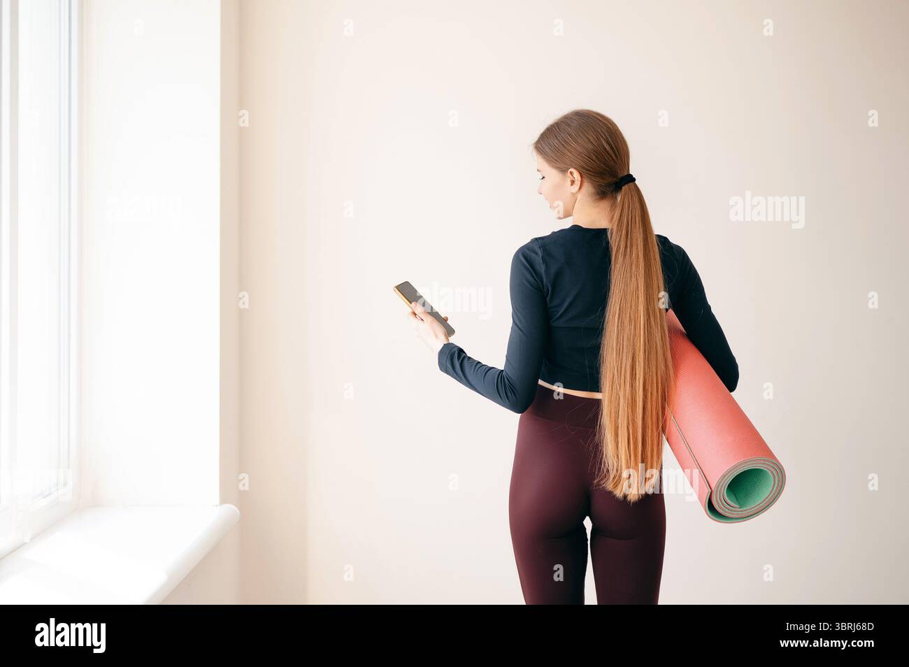 Frau mit Pferdeschwanz, die beim Training oder Yoga-Kurs stand, mit einer rollenden Fitnessmatte und einem Smartphone in ihrem Mann drehte sie der Kamera den Rücken zu. Speicherplatz kopieren auf Stockfoto